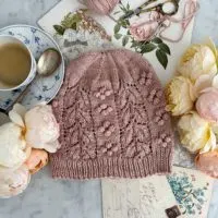 A Top-down view of the Laurel Hat, a pink hand knit hat with bobbles and lacy leaves, which is laid flat on a white marble countertop. It's surrounded by roses, a teacup full of espresso, and antique paper ephemera.