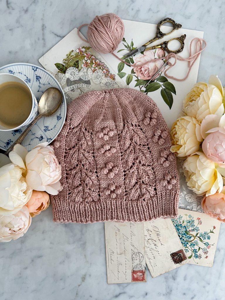 A Top-down view of the Laurel Hat, a pink hand knit hat with bobbles and lacy leaves, which is laid flat on a white marble countertop. It's surrounded by roses, a teacup full of espresso, and antique paper ephemera.