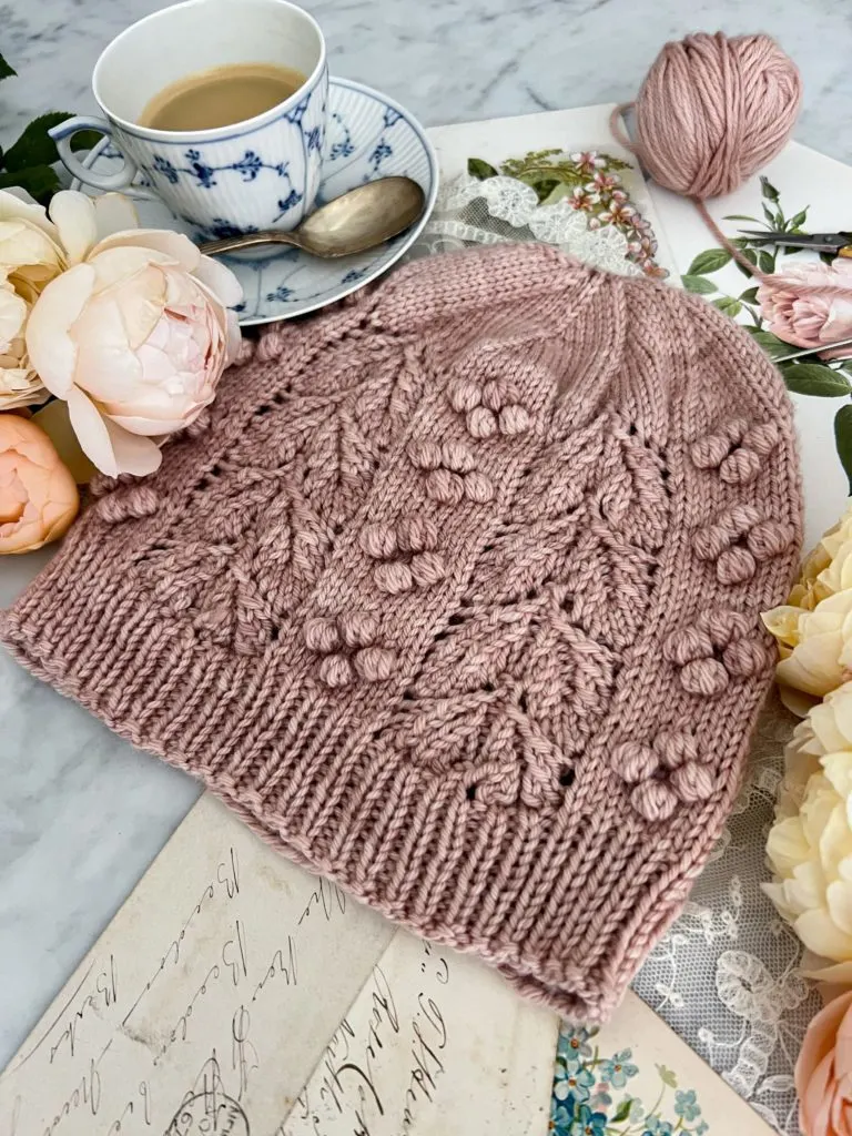 An angled image of the Laurel Hat, a pink hand knit hat with bobbles and lacy leaves. The image is taken from the right-hand corner of the brim upward toward the crown.