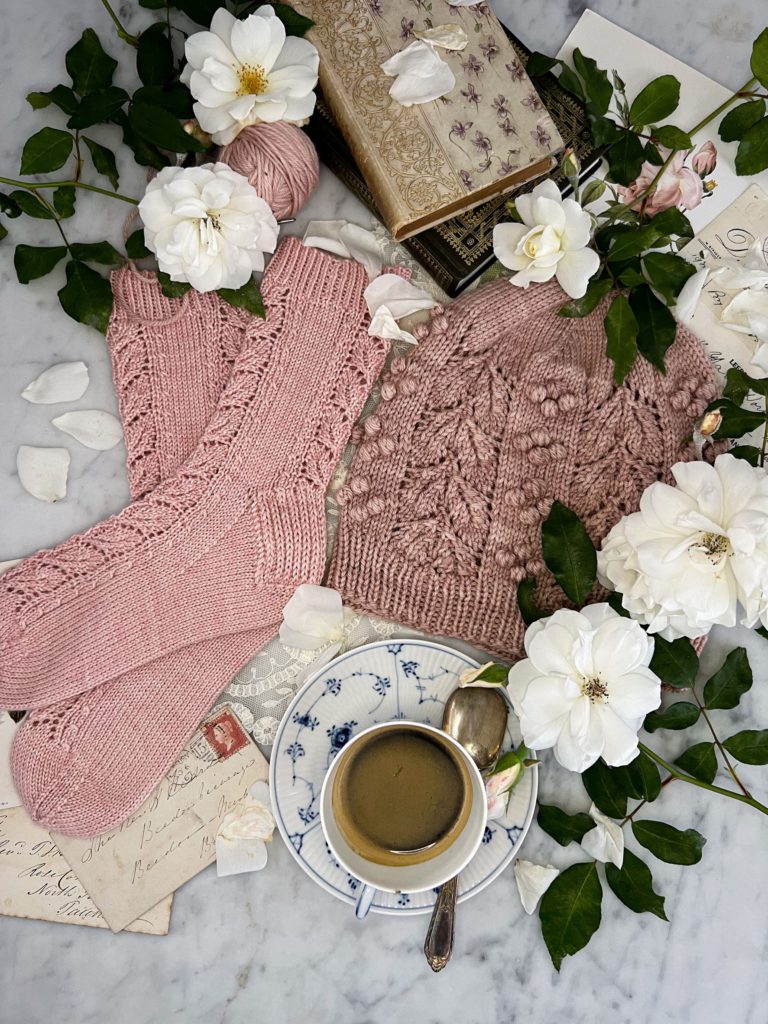 A top down photo of a pair of pink knit socks and a pink knit hat. They're surrounded by white roses, antique books, and a teacup full of espresso.
