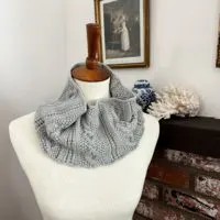 A gray, cabled cowl is looped around the neck of a white dressmaker's form. In the background is a brick fireplace with coral and a ginger jar on the mantel, along with some antique prints on the wall.