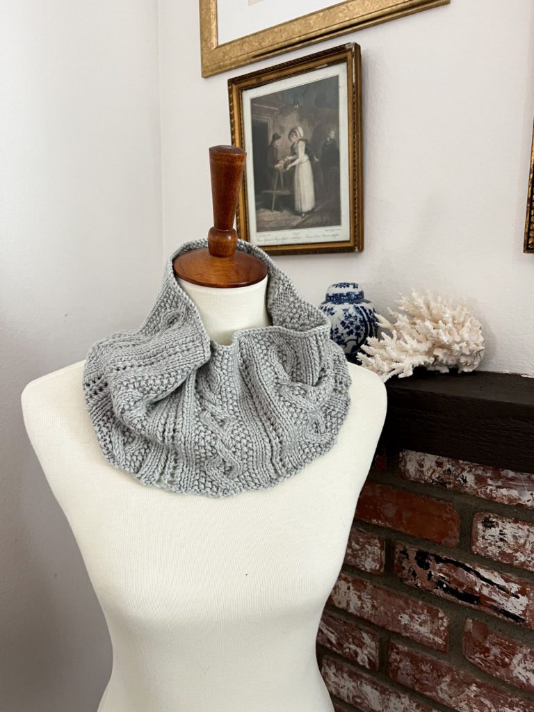 A gray, cabled cowl is looped around the neck of a white dressmaker's form. In the background is a brick fireplace with coral and a ginger jar on the mantel, along with some antique prints on the wall.