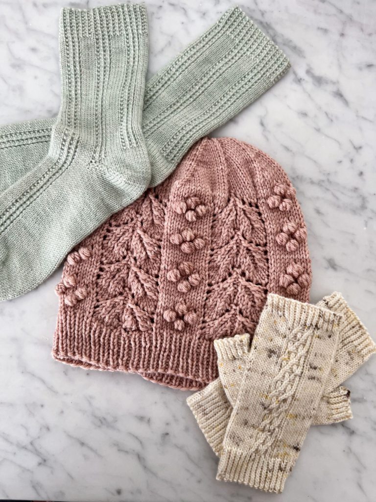 A pair of seafoam green knit socks in the top left, a pink knit hat in the middle, and a pair of cream handknit mitts in the bottom right
