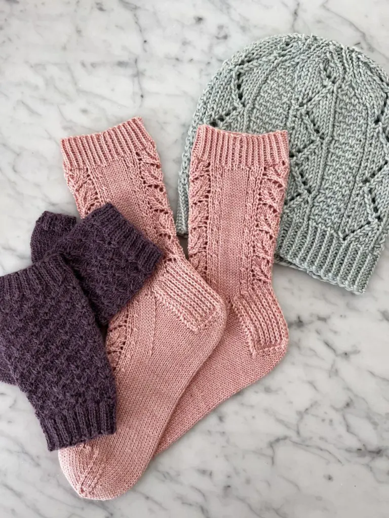 A pair of purple handknit mitts on the bottom left, a pair of pink socks in the middle, and a light blue knit hat in the top right