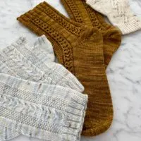 A pair of light blue variegated knit mitts in the foreground with a pair of caramel-colored knit socks in the middle and a pair of cream knit mitts in the very back