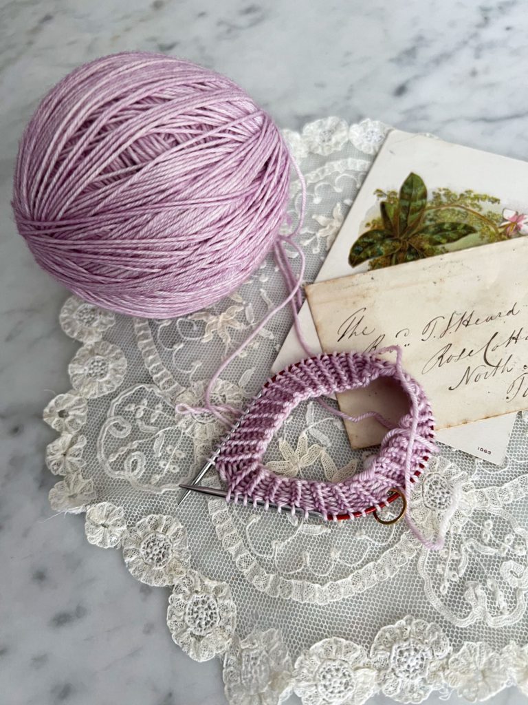 A top-down image of the beginnings of a lavender-colored knit sock, a lavender-colored ball of yarn, and an antique envelope and postcard, all sitting on a white lace doily.