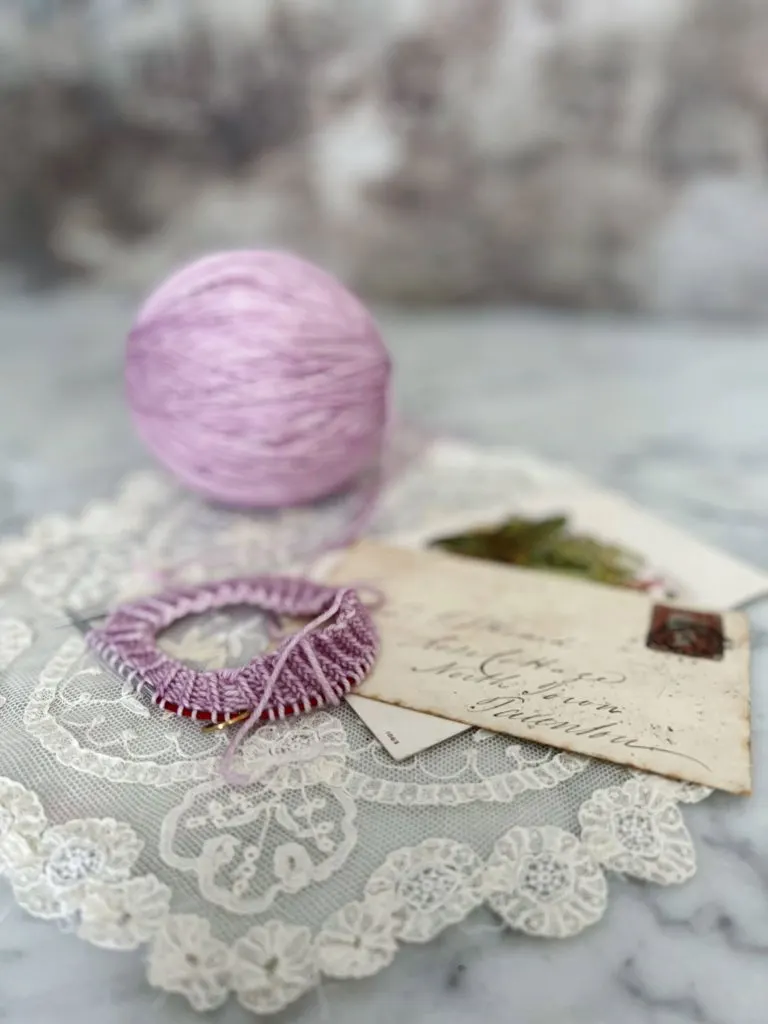 The beginnings of a lavender-colored knit sock sit in the foreground on a white lace doily. Blurred in the background are a ball of yarn and some antique paper ephemera.