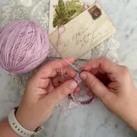 A woman's small, plump, white hands work on knitting the beginnings of a lavender hand-knit sock.