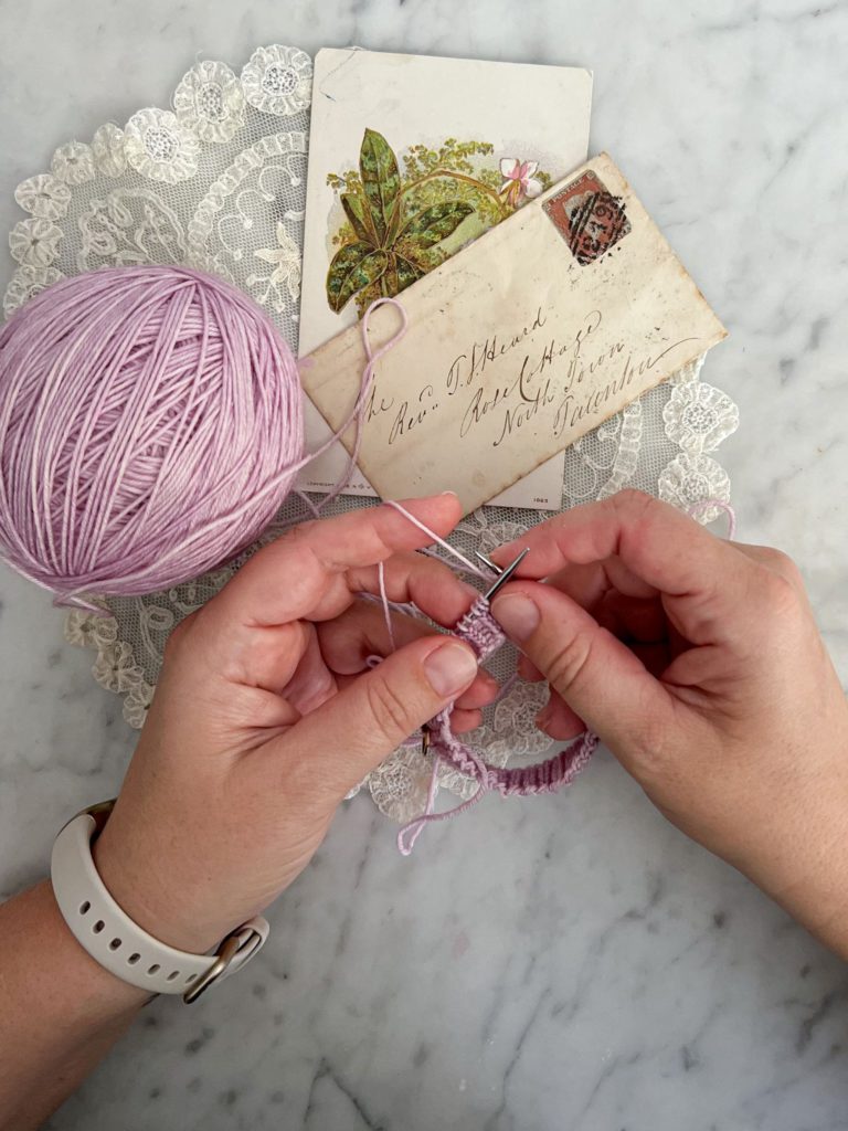A woman's small, plump, white hands work on knitting the beginnings of a lavender hand-knit sock.
