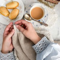 A top-down photo of a woman's pair of small, plump, white hands knitting a seed stitch shawl with a mohair edging. There's a cup of milky tea and a plate of madeleine cookies nearby.