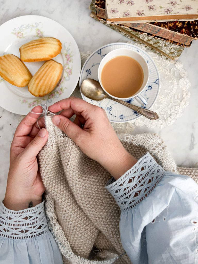 A top-down photo of a woman's pair of small, plump, white hands knitting a seed stitch shawl with a mohair edging. There's a cup of milky tea and a plate of madeleine cookies nearby.