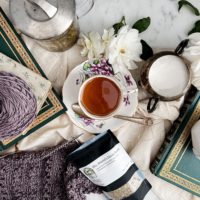 A top-down image a flatlay on a white marble countertop. In the center is a teacup with golden tea on a saucer with purple flowers painted on it. They're surrounded by a glass teapot, white roses, antique books, a sugar bowl, a bag of loose leaf tea, and a purple sweater in progress on two steel knitting needles.