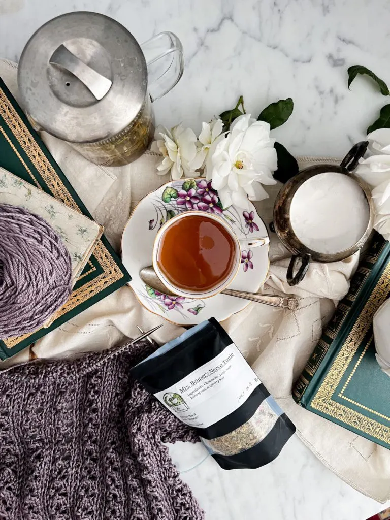 A top-down image a flatlay on a white marble countertop. In the center is a teacup with golden tea on a saucer with purple flowers painted on it. They're surrounded by a glass teapot, white roses, antique books, a sugar bowl, a bag of loose leaf tea, and a purple sweater in progress on two steel knitting needles.