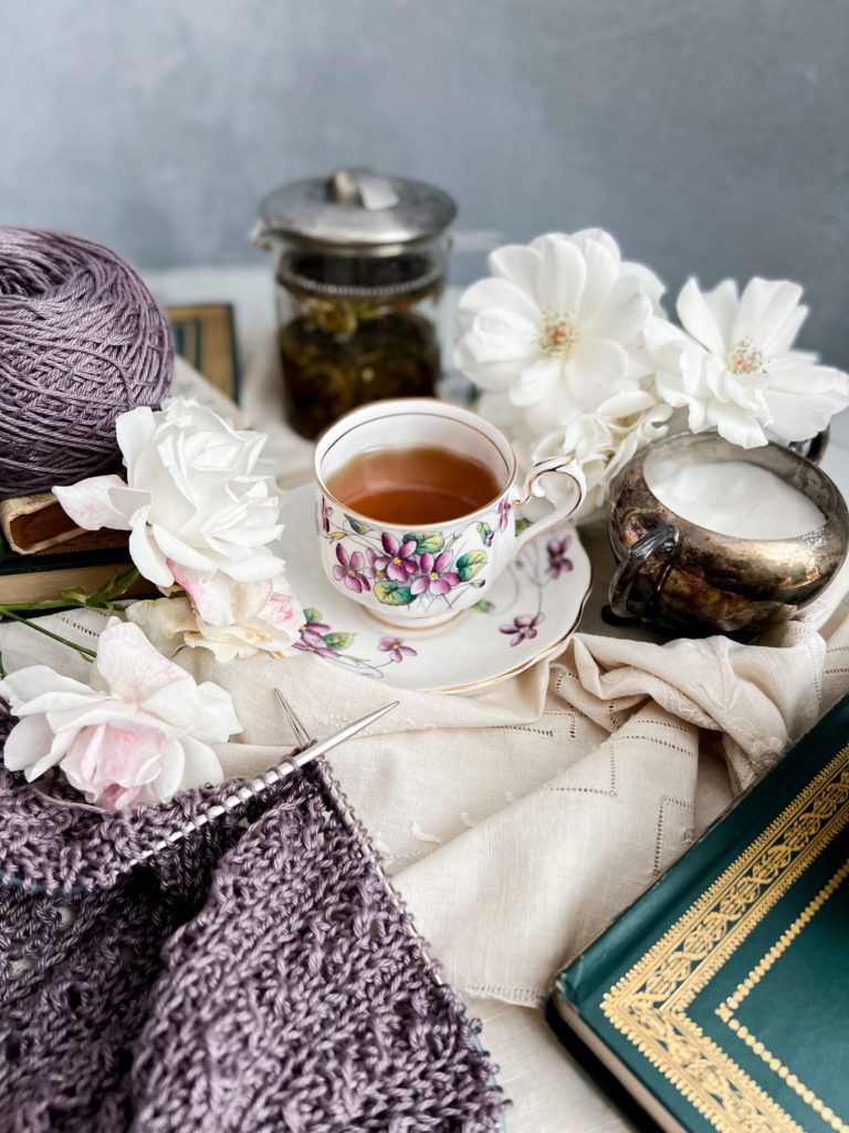 A teacup with purple flowers sits in the center of the photo, surrounded by white roses, a purple sweater in progress on two steel knitting needles, and some antique books.