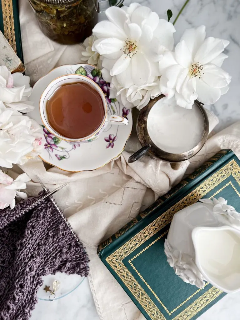A top down image of a teacup filled with golden tea. The teacup is in the top left of the photo. Around it are white roses, a green book with gilt trim on the cover, antique linens, and a purple sweater in progress on two steel needles.