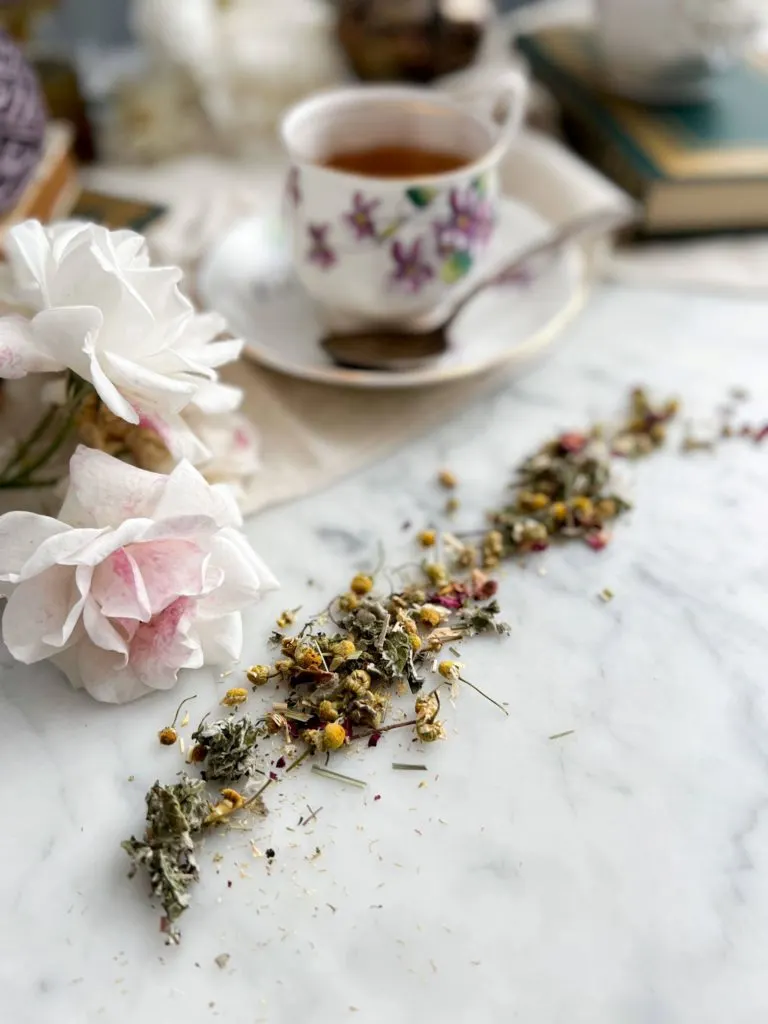 In the foreground, a bit of tea with chamomile, lemon grass, and rose petals is spread on a white marble countertop. Blurred in the background are white roses, a teacup full of tea, and some antique books.