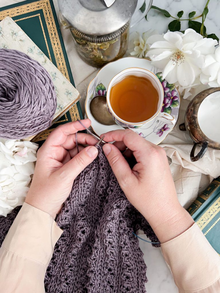 A top-down photo of a woman's pair of small, plump, white hands knitting a purple cotton sweater. Nearby are white roses, antique books, and a teacup with purple flowers filled with light golden tea.