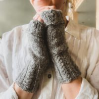 A pair of gray fingerless mitts with cabled cuffs on a woman's hands clasped together in front of her chin.
