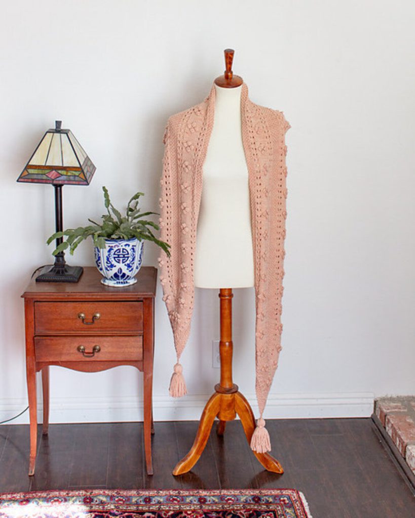 A pink scarf with pointed ends is draped loosely around the shoulders of a white dressmaker's form. The ends are dangling down to the floor, and each point is adorned with a tassel. To the left is a small antique table with a potted plant and a stained glass lamp on it.