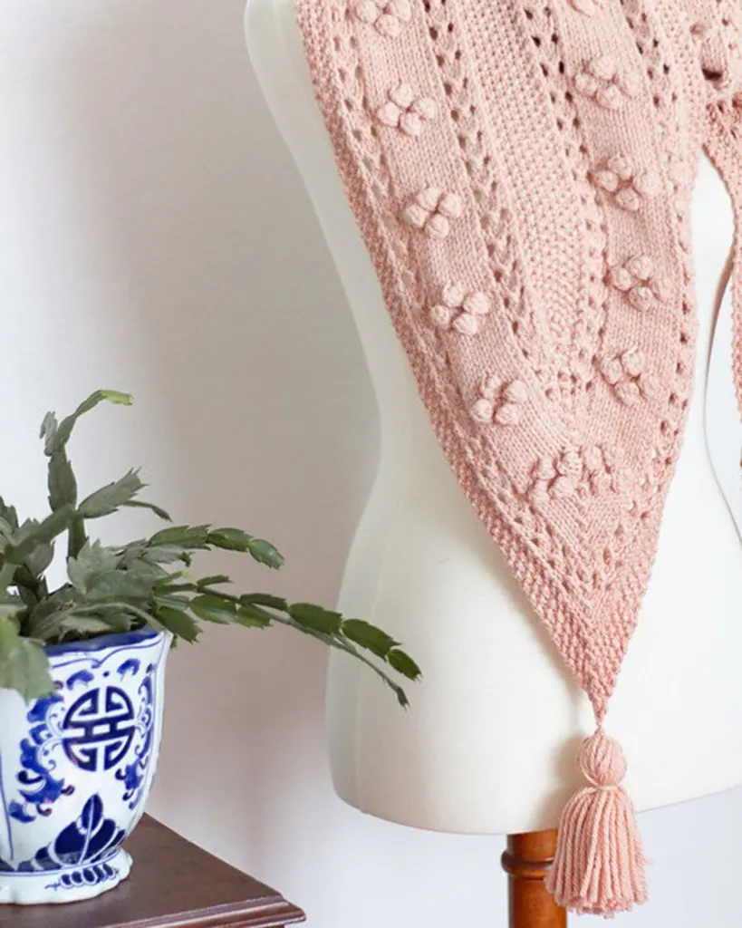 A close-up on one of the pointed ends of the Miramar Scarf, a pink knit scarf with eyelets, seed stitch, and bobbles. A tassel hangs from the end of the point.