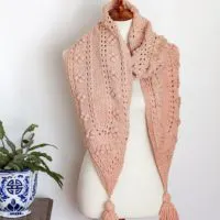 The Miramar Scarf, a pink knit scarf with pointed ends, is wrapped around the neck of a white dressmaker's form. Each end of the scarf comes to a point and is decorated with a tassel.