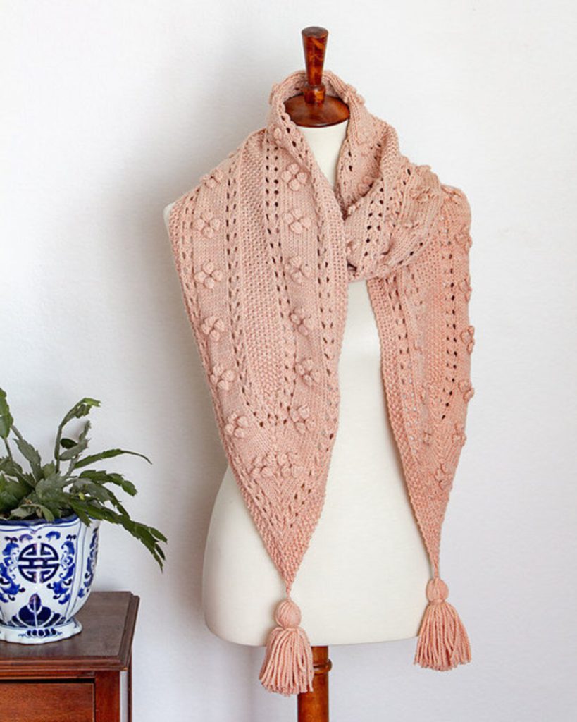 The Miramar Scarf, a pink knit scarf with pointed ends, is wrapped around the neck of a white dressmaker's form. Each end of the scarf comes to a point and is decorated with a tassel.