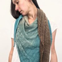 A woman with long brown hair wears a shawl draped around her shoulders. She is looking to the left and downward so you can see the shawl's drape. The shawl is knit in a blue and brown gradient fade yarn with vertical panels of seed stitch and lace.