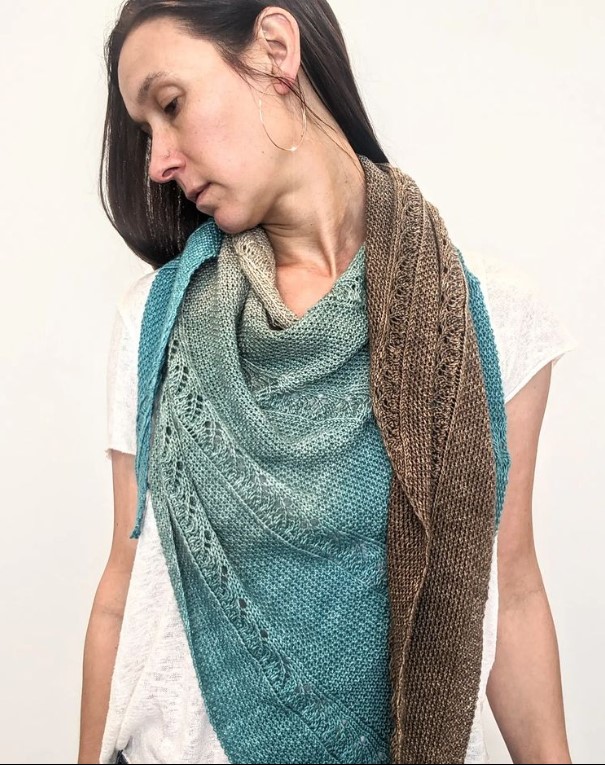A woman with long brown hair wears a shawl draped around her shoulders. She is looking to the left and downward so you can see the shawl's drape. The shawl is knit in a blue and brown gradient fade yarn with vertical panels of seed stitch and lace.