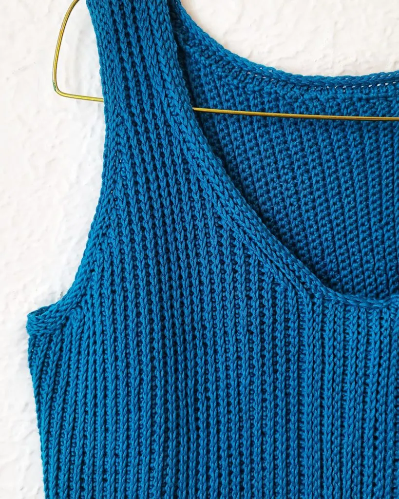 A rich blue tanktop hangs from a wire hanger against a white stucco wall. The tanktop is crocheted, and the image is zoomed in on the right shoulder and neckline shaping.