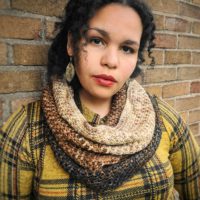 A brown woman with black hair is standing against a brick wall. She is wearing a yellow plaid shirt while looking straight at the camera with her head slightly cocked to the side; a brown cowl is draped around her neck.