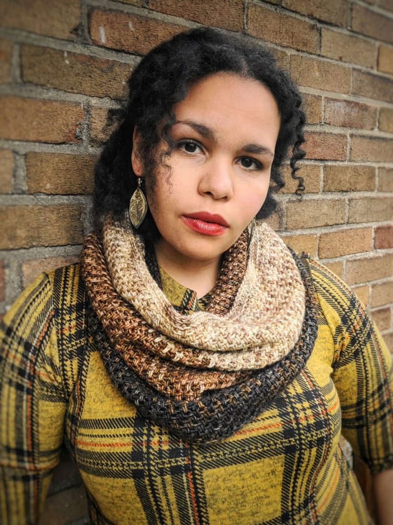 A brown woman with black hair is standing against a brick wall. She is wearing a yellow plaid shirt while looking straight at the camera with her head slightly cocked to the side; a brown cowl is draped around her neck.