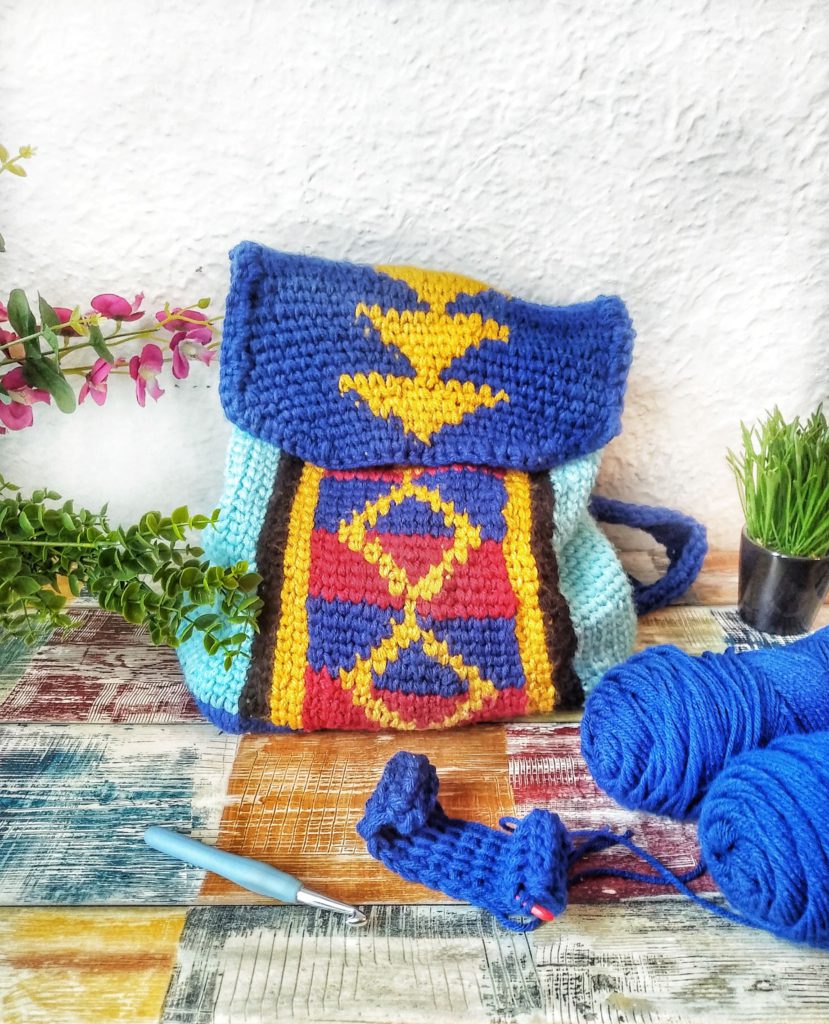 A colorful crocheted backpack in shades of blue and aqua with center panels of yellow diamonds on blue and red stripes sits on the floor against a white stucco wall. In the foreground are some skeins of blue yarn, a crochet hook, and a small crochet WIP.