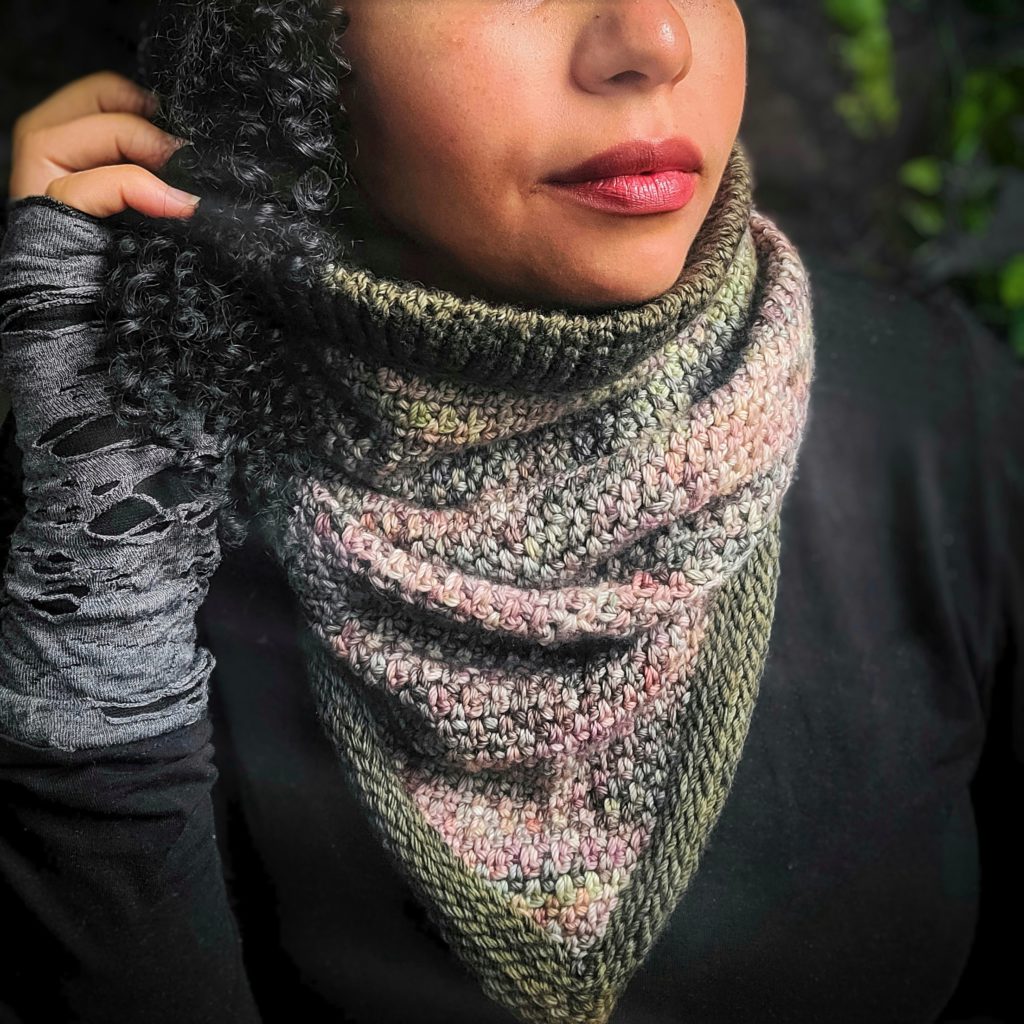A brown woman with black hair wears a bandana-shaped cowl around her neck. The cowl is crocheted with yarn in colors that echo a softly faded bouquet of hydrangeas. Only the lower 2/3 of the woman's face is visible.