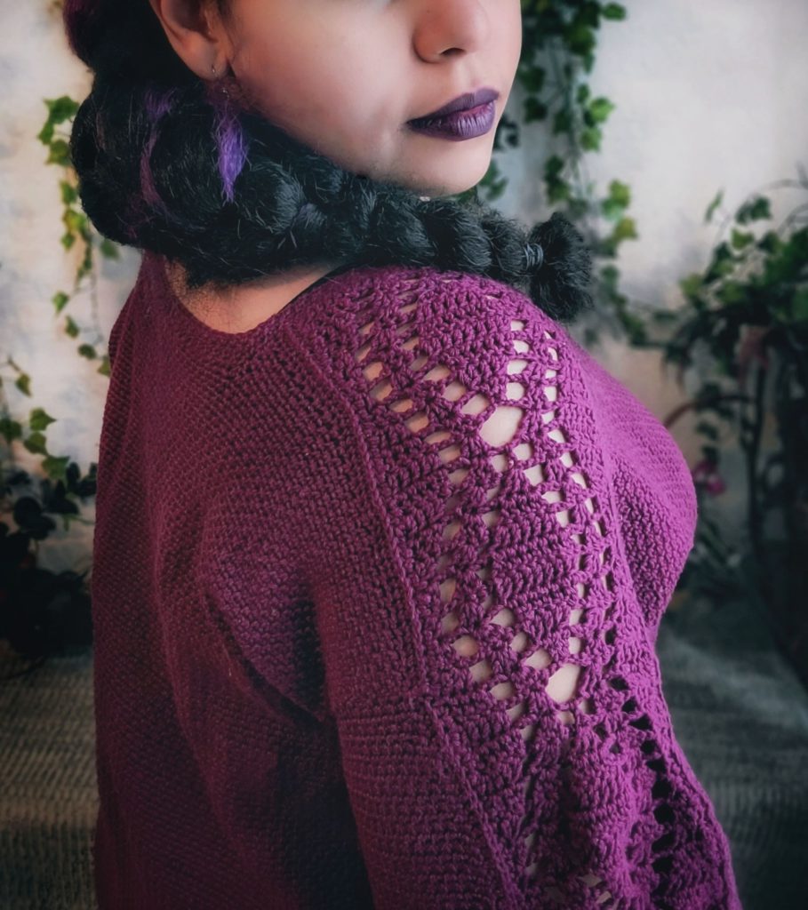 A brown woman with black hair in a long braid to the side wears a purple crocheted sweater. The sweater has a plain body with lady sleeves. The woman is turned to the side to show the sleeve detail.