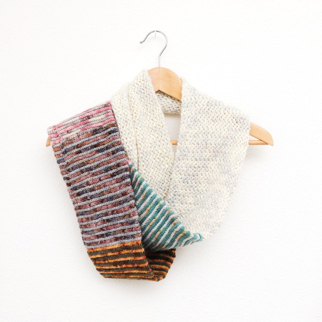 A crocheted cowl is draped around a wooden hanger suspended from a nail on the wall. The cowl is half colorful stripes, half softly-speckled cream and gray.