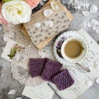 A top-down image of three purple yarn swatches, a teacup full of espresso, some old books, and a pitcher full of roses just peeking into the left corner of the image.