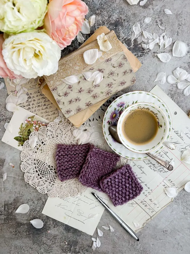 A top-down image of three purple yarn swatches, a teacup full of espresso, some old books, and a pitcher full of roses just peeking into the left corner of the image.