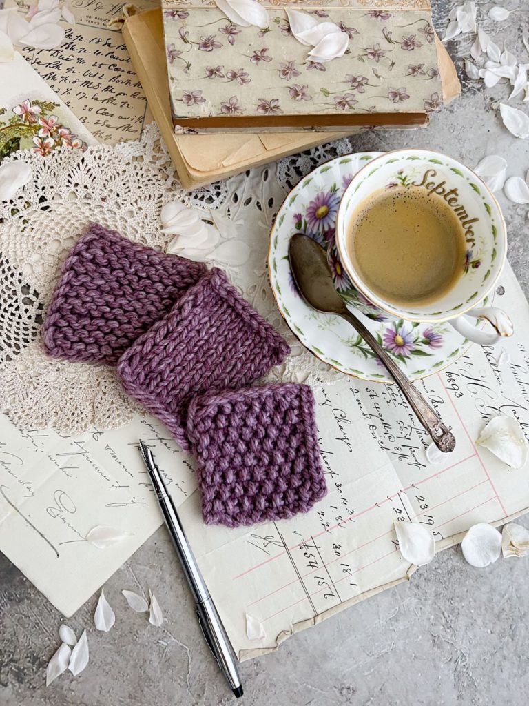 Three purple swatches are arranged in a soft arc in the bottom left of the image. To the right and above are a teacup full of espresso and some vintage books.