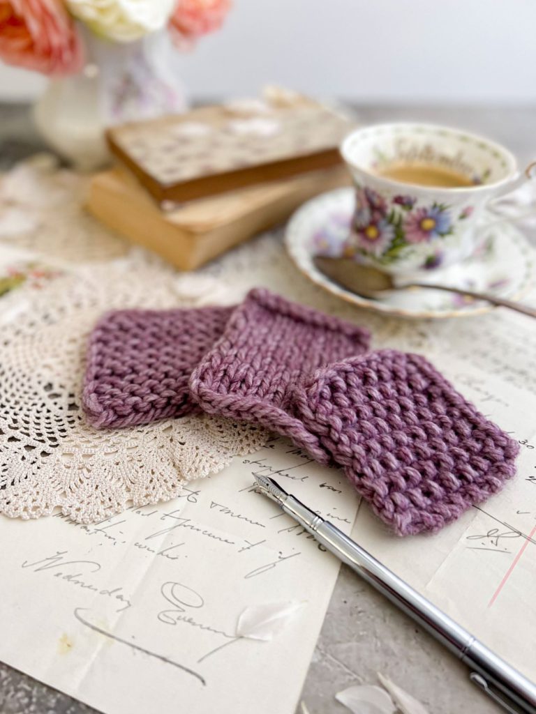Three purple swatches are arranged in a soft arc in the foreground on top of some antique paper ephemera and a doily. Blurred in the background are a teacup full of espresso, some old books, and roses in a pitcher.