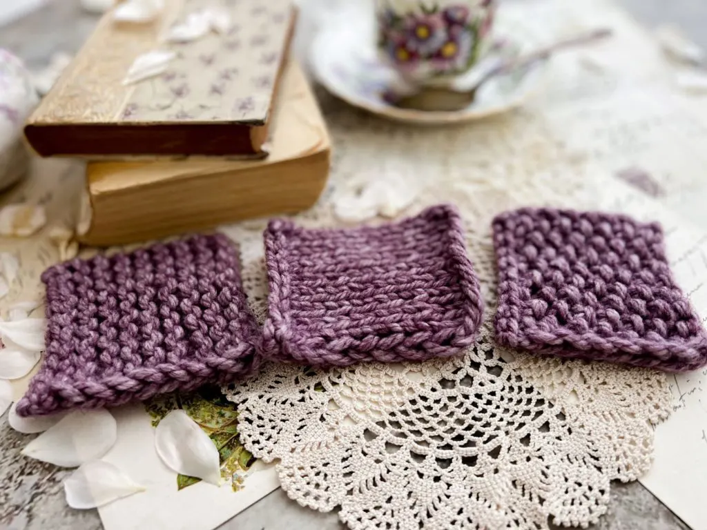 Three purple yarn swatches are laid out in a row. On the left is garter stitch, in the middle is stockinette, and on the right is seed stitch. The edges that have a slip stitch edging are facing toward the camera.