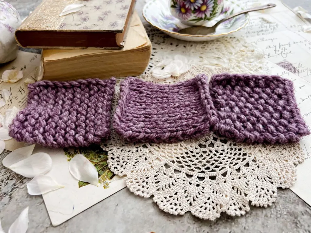 Three purple yarn swatches are laid out in a row. On the left is garter stitch, in the middle is stockinette, and on the right is seed stitch. The edges that don't have a slip stitch edging are facing toward the camera.