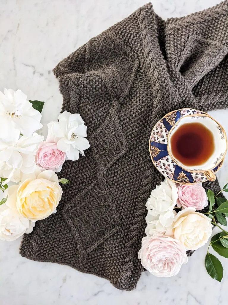 A brown cabled scarf is laid flat on a white marble countertop. It is surrounded by pink, white, and yellow roses and a teacup full of Earl Grey.