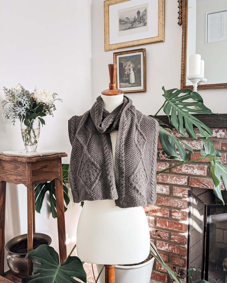 A brown cabled scarf is wrapped around the neck of a white dressmaker's form with the ends dangling down the front. In the background are a brick fireplace, some house plants, and framed antique prints on the wall.