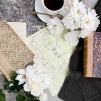 A top-down image of a design for a cabled scarf (in the very center of the photo) surrounded by antique books, white roses, a white teacup and saucer, and a little glimpse of the finished scarf made from the design.