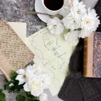 A top-down image of a design for a cabled scarf (in the very center of the photo) surrounded by antique books, white roses, a white teacup and saucer, and a little glimpse of the finished scarf made from the design.