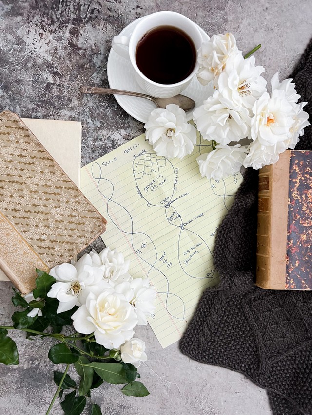 A top-down image of a design for a cabled scarf (in the very center of the photo) surrounded by antique books, white roses, a white teacup and saucer, and a little glimpse of the finished scarf made from the design.