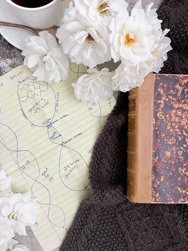 A close-up image of the sketch for a cabled scarf that takes up the left 2/3 of the photo. On the right is a little glimpse of the finished scarf with an old book on it. Up top are white roses and a white teacup with saucer.