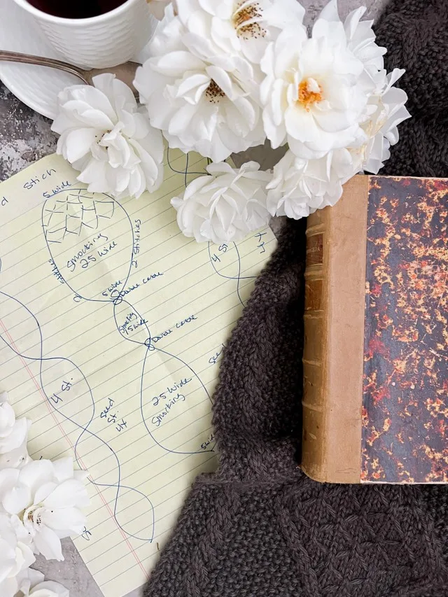 A close-up image of the sketch for a cabled scarf that takes up the left 2/3 of the photo. On the right is a little glimpse of the finished scarf with an old book on it. Up top are white roses and a white teacup with saucer.