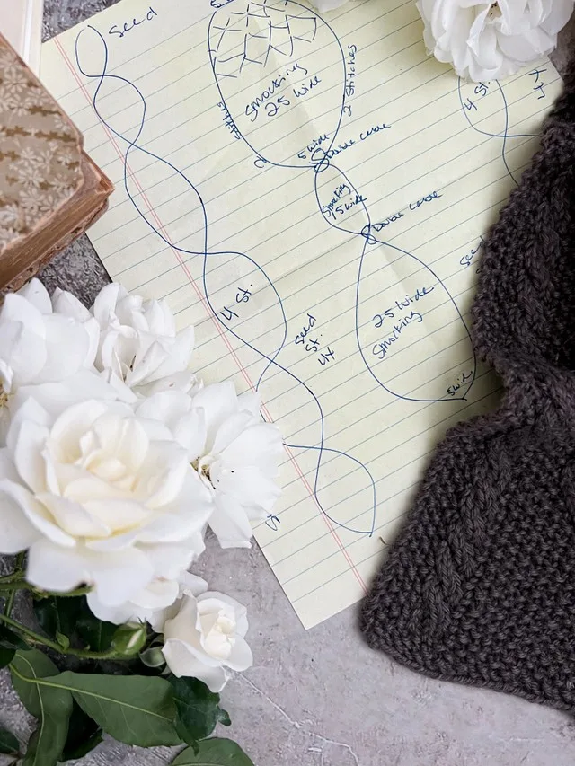 A zoomed-in image of a sketch for a cabled scarf. Around it are white roses, an old book, and a tiny peek at a corner of the finished scarf.