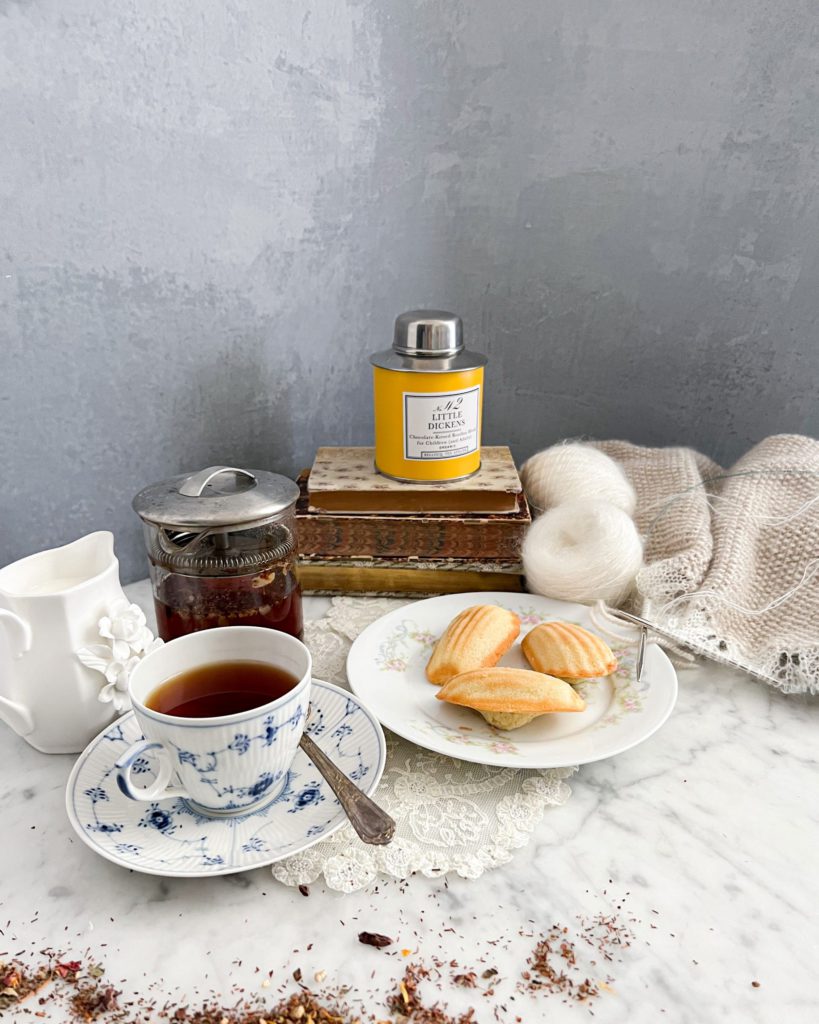 A yellow canister of tea sits on top of a tall stack of books surrounded by knitting, madeleines, a clear pot of tea, a creamer, and a blue and white teacup filled with tea.